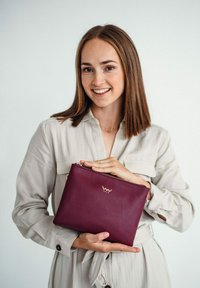 Burgundská kožená kabelka přes rameno se zlatým logem. Zipové uzavírání, obdélníkový design, hladký povrch, držena osobou v světlé košili.