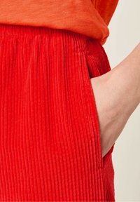 Pantalons en velours côtelé rouge avec des côtes verticales, dotés d'une poche latérale et d'une taille élastique. La texture douce renforce l'attrait visuel.