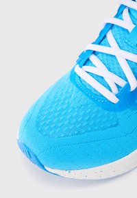 Zapatilla deportiva azul claro con parte superior de tela texturizada, cordones blancos y media suela blanca con moteado. Forma angular con diseño aerodinámico.