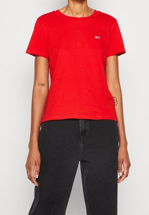 Person wearing a plain red short-sleeve shirt with small logo on chest and high-waisted black jeans, showing arm tattoos.