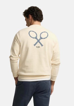 Hombre con sudadera cremosa con gráfico de tijeras azules cruzadas y texto "SLBN" en la espalda, combinado con pantalones azul oscuro.