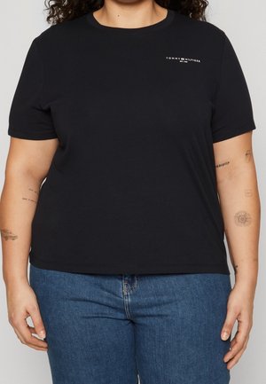 Osoba ubrana w czarną koszulkę Tommy Hilfiger i niebieskie dżinsy, pokazująca tatuaże na obu przedramionach na neutralnym tle.