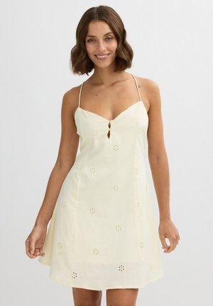 Mujer con cabello castaño hasta los hombros, que lleva un vestido sin mangas color crema con patrones florales calados y tirantes delgados, sonriendo a la cámara.