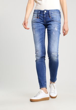 Schlanke blaue Denim-Jeans mit distressed Details und Ausbleichungen. Fünf-Taschen-Design mit einem vorderen Knopfverschluss, kombiniert mit weißen Sneakers.