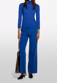 Mörkblå åtsittande långärmad topp med hög krage, i kombination med löst sittande mörkblå byxor. Svarta spetsiga mules och en silverfärgad väska.