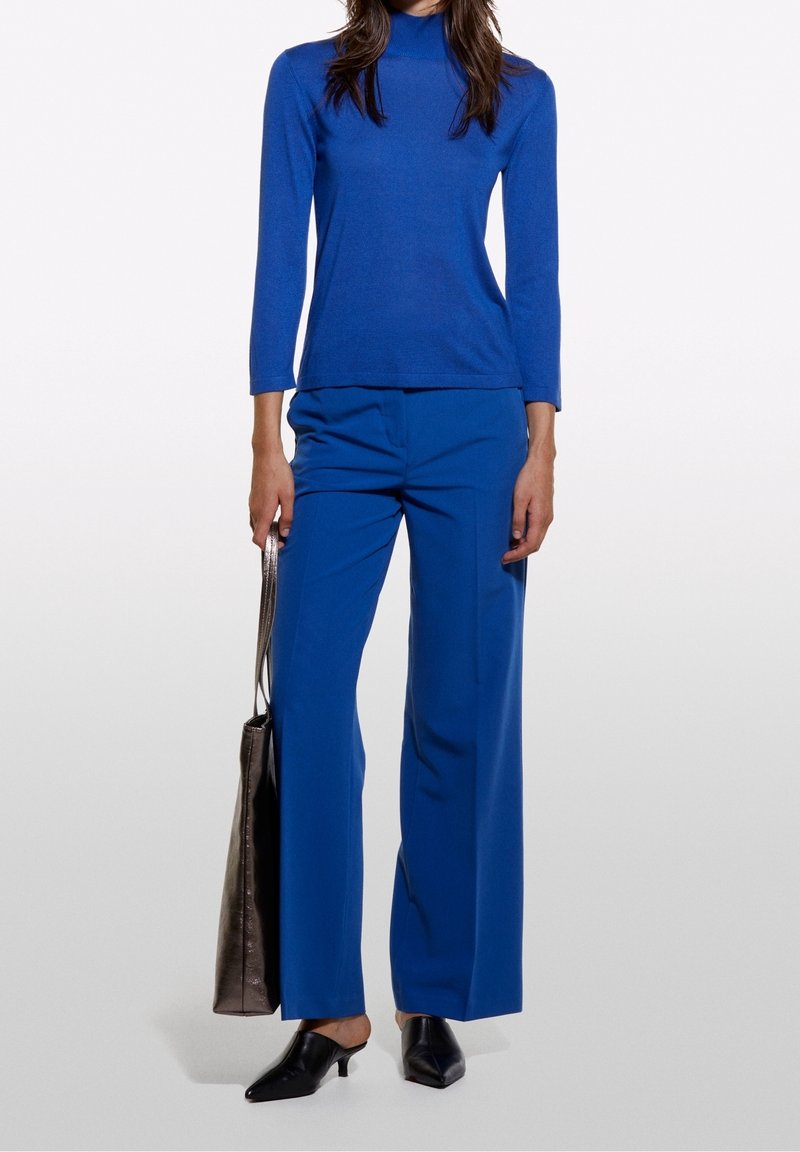 Mörkblå åtsittande långärmad topp med hög krage, i kombination med löst sittande mörkblå byxor. Svarta spetsiga mules och en silverfärgad väska.