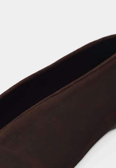 Gros plan d'une chaussure à enfiler marron foncé avec une tige en tissu doux et une ouverture légèrement incurvée sur fond blanc.