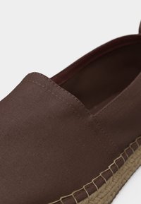 Chaussure en toile marron à enfiler avec coutures visibles et semelle en jute tissée, gros plan sur fond blanc.