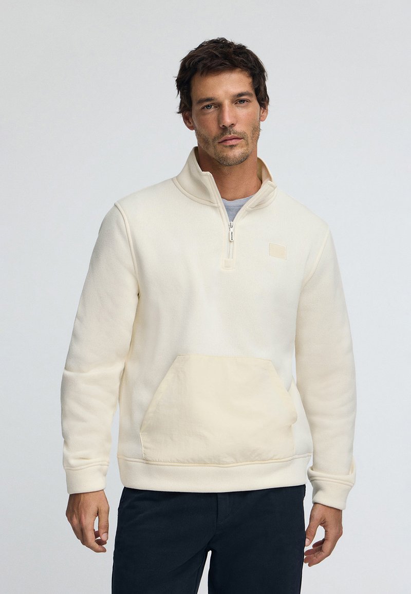 Sudadera de color crema con cuello de media cremallera, bolsillo frontal y textura suave. Presenta un discreto parche con el logo en el pecho.