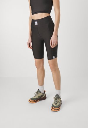 Osoba nosící černé cyklistické šortky s vysokým pasem a bočními kapsami, černý crop top, bílé ponožky a zeleno-oranžové trailové běžecké boty.