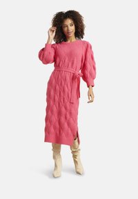 Donna con capelli ricci che indossa un vestito midi rosa in maglia con cintura, maniche a sbuffo e stivali crema al ginocchio, in piedi su uno sfondo bianco.