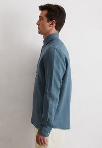 Langärmliges Button-Up-Hemd in einem verblassten Teal, aus strukturiertem Stoff, mit einem geraden Kragen und weißen Knöpfen, seitlich gezeigt.