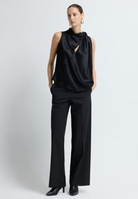 Zwarte satijnen mouwloze top met gedrapeerde halslijn, gecombineerd met hoge zwarte broeken en puntige hakken, staand tegen een lichte achtergrond.