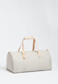 Borsa da viaggio in canvas beige con manici in pelle color sabbia, forma arrotondata, zip dorata e cuciture a contrasto. Nessun motivo o logo visibile.