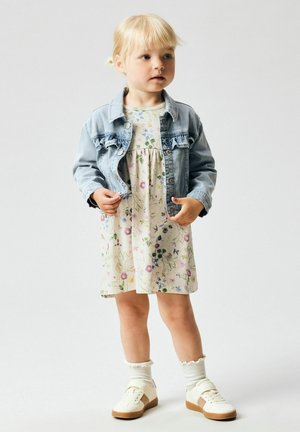 Jong meisje dat een bloemenjurk draagt, een licht denim jack, witte sokken en witte sneakers, staand tegen een effen lichte achtergrond.