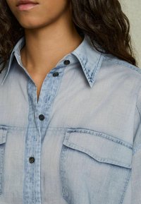 Camicia di denim azzurro chiaro con due tasche sul petto, colletto a camicia e bottoni neri. Presenta una consistenza morbida e dettagli di scolorimento sottili.