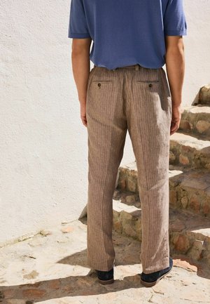 Férfi áll köves lépcsőfokon, barna csíkos nadrágot és kék rövid ujjú inget visel, és egy fehér, texturált falnak háttal áll.