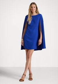 Rövid, testhez simuló kék ruha cape ujjakkal. Simább szövet egyenletes színnel és kerek nyakkivágással. Barna sarokkal ellátott szandálok.