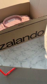 Cardboard box labeled "Zalando" containing a folded striped shirt in pink and white. Visible on a marble surface with a red table leg.