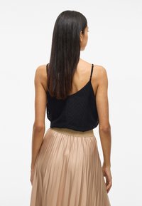 Čierna textúrovaná tielko so zigzagovým vzorom; skombinované s plisovanou, vysoko pásovou, svetlobežovou sukňou. Zadný pohľad ukazuje remienky a vlasy.