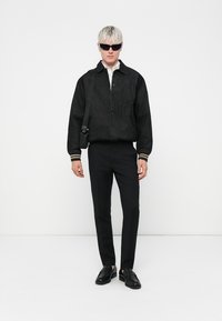 Svart denimjakke med knappeåpning, ribbestrikkede mansjetter, kombinert med skreddersydde svarte bukser og svarte loafers; modell iført solbriller.