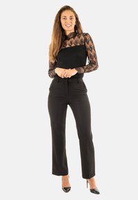Zwarte blouse met lange mouwen en kantdetails, gecombineerd met zwarte high-waisted broek. Model staat met de handen gevouwen, draagt zwarte hakken.