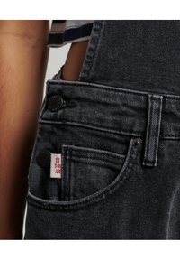 Donkere denim salopettes met een textuurafwerking, voorzien van messing knopen, een voorzak met een wit label, en zijopeningen voor verstelbaarheid.