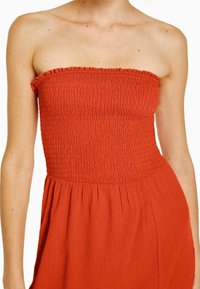 Off-shoulder klänning i bränd orange, med smockad överdel, texturerad tyg och flödande kjol med subtila veck.