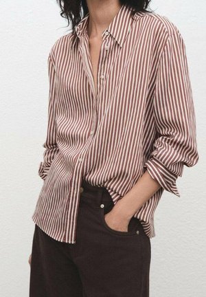 Femme portant une chemise à rayures verticales marron et blanches avec les manches retroussées, associée à un pantalon foncé, main dans la poche.