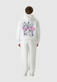 Hvit hettegenser med grafisk trykk på ryggen som viser skisseaktige bilder og rosa tekst "lost youth." Kombinert med matchende hvite joggebukser.