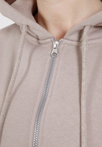 Felpa beige con zip, con zip in metallo, lacci e tessuto testurizzato. Presenta un cappuccio accogliente e dettagli di cucitura visibili.