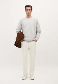 Sweat-shirt gris clair, pantalon crème, veste en daim marron portée sur le bras, baskets blanches avec des accents bleus. Look décontracté, coupe ajustée.