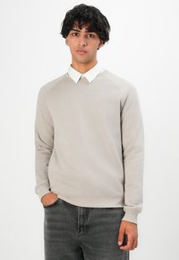 Jonge man met donker krullend haar draagt een lichtbeige sweatshirt over een witte shirt met kraag en donkergrijze spijkerbroek, staand met één hand in zijn zak.