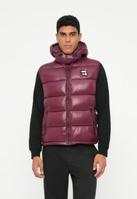 Bordo puffer prsluk s visokim ovratnikom, prednjim patentnim zatvaračem i dva bočna džepa. Sadrži logotip na lijevoj strani prsiju i sjajnu teksturu.