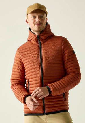 Mann trägt eine orange gesteppte Jacke und eine beige Kappe, zieht den Ärmel hoch, leicht nach rechts gewandt vor einfarbig hellem Hintergrund.