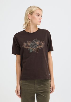 Lyshåret kvinde iført en mørkebrun T-shirt med blomster- og vingedesign, kombineret med olivengrønne bukser, kigger mod venstre.