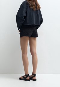 Czarna krótka kurtka, czarne postrzępione krótkie spodenki oraz czarne sandały na paski. Strój cechuje się swobodnym krojem i minimalistycznymi akcentami w postaci metalowych detali.