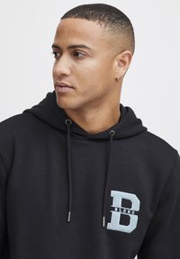 Sudadera negra con capucha ajustable, que presenta un logo frontal "B L E N D" en azul claro texturizado. Tejido suave, diseño relajado.
