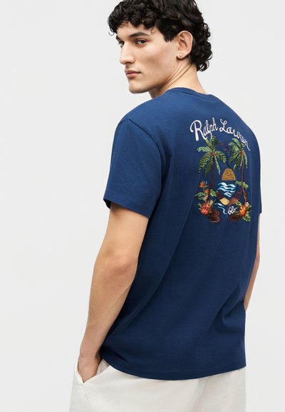 Νέος άνδρας με σγουρά σκούρα μαλλιά που φοράει μπλε ναυτικό t-shirt Ralph Lauren με κεντητό τροπικό σχέδιο στην πλάτη.