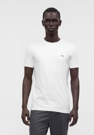 Jeune homme aux cheveux courts portant un t-shirt blanc uni à manches courtes et un pantalon gris, debout devant un fond clair uni.