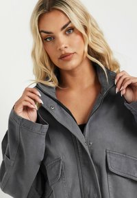 Chaqueta gris con cremallera frontal y un acabado texturizado, con grandes bolsillos y un cuello alto, usada parcialmente desabrochada, mostrando un ajuste casual.