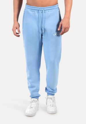Hellblaue Baumwollsweatpants mit verstellbarem Kordelzug, seitlichen Taschen und gesticktem Logo. Kombiniert mit weißen Sportschuhen.