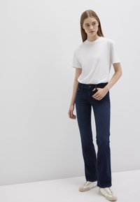 Flared dark blue jeans worn with a white t-shirt, featuring a fitted waistband and subtle stitching details, paired with light-colored sneakers.