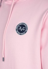 Světle růžová mikina s kapucí a kruhovým námořnickým logem s písmenem V a textem. Obsahuje šňůrku s kovovým hrotem. Hladký design textury.