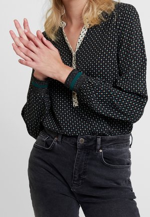 Vrouw met een blouse met lange mouwen en dessin, voorzien van contrasterende manchetten en kraag, gecombineerd met een hoge taille zwarte spijkerbroek, handen samen gevouwen.