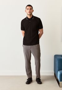 Zwart poloshirt met een gestructureerd patroon, korte mouwen en witte kraagaccenten, gecombineerd met grijze broek en zwarte sneakers.