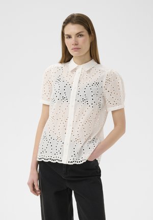 Junge Frau trägt eine weiße Bluse mit kurzen Ärmeln aus Augenschlitzstoff, schwarzer Unterlage und schwarze Hose, steht mit einer Hand in der Tasche.