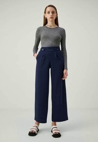 Grå ribbestrikket langermet topp kombinert med marineblå vide bukser med høy midje, som har frontlommer og knappedetaljer, brukt med svarte og hvite sandaler.