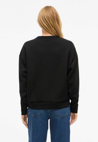 Woman with long blonde hair wearing a plain black sweatshirt and blue jeans, viewed from behind against a white background.