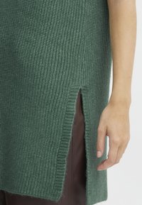 Grüne gestrickte Tunika mit gerippter Textur, seitlichem Schlitzdetail und lockerer Passform. Der Stoff wirkt weich und dick, was zu einer braunen Unterlage passt.